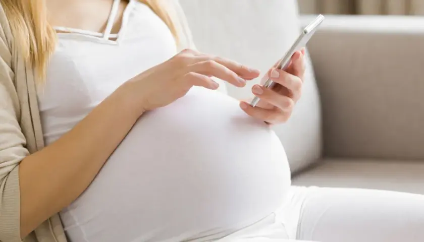 Kobieta w 24. tygodniu ciąży (l4 w ciąży) przegląda coś na telefonie, siedząc na kanapie.