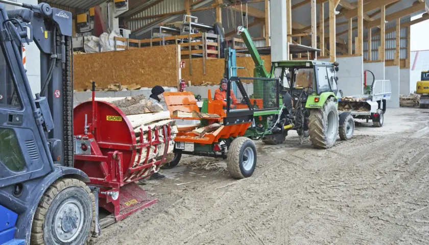 Ein Gabelstapler lädt Holz auf einen Tischholzspalter. Ein Traktor mit angehängtem Wagen wartet daneben.