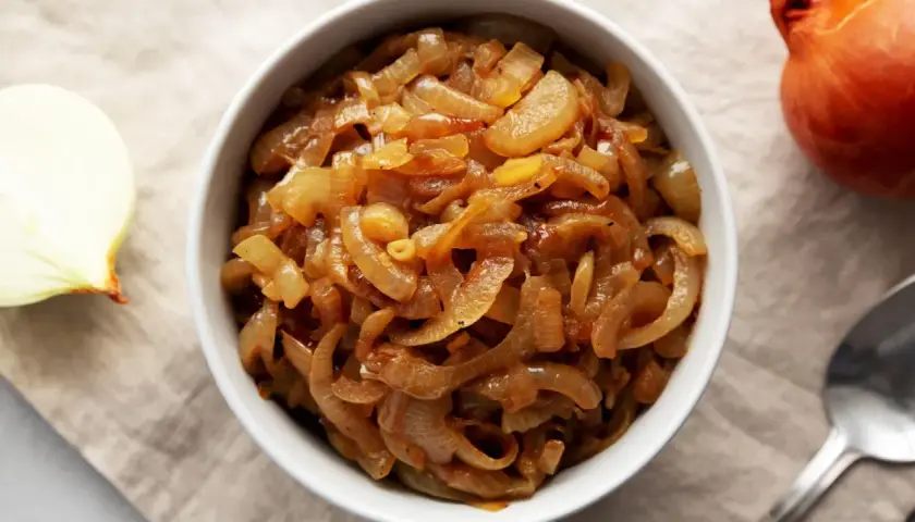 Un bol rempli d'oignons caramélisés dorés, parfaits pour une recette savoureuse. Un oignon cru à côté.