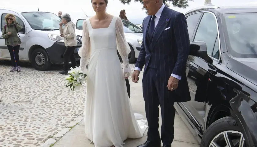 Novia con ramo de lirios y su acompañante, un hombre con traje de raya diplomática, llegan a su boda. Irene Matamoros, radiante.