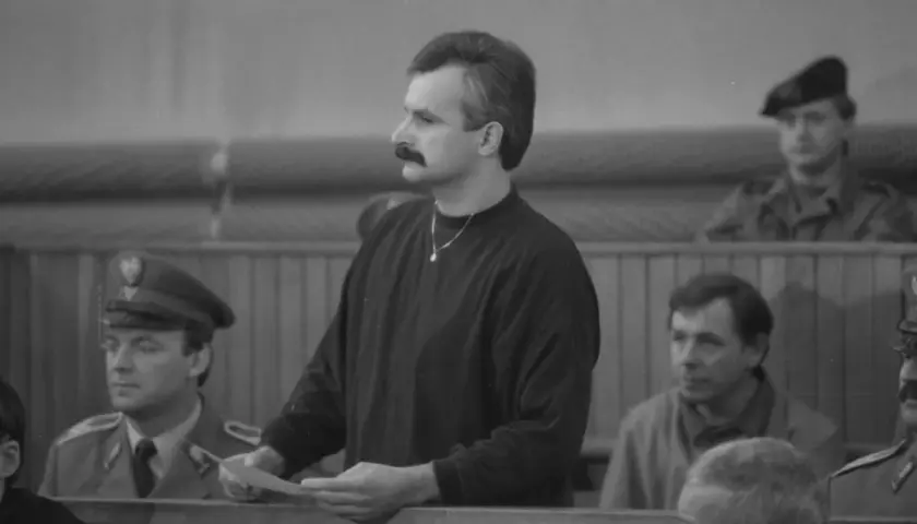 Mężczyzna z wąsami, niczym minstrel, trzyma dokumenty w sali sądowej. Obok niego siedzą ludzie w mundurach.