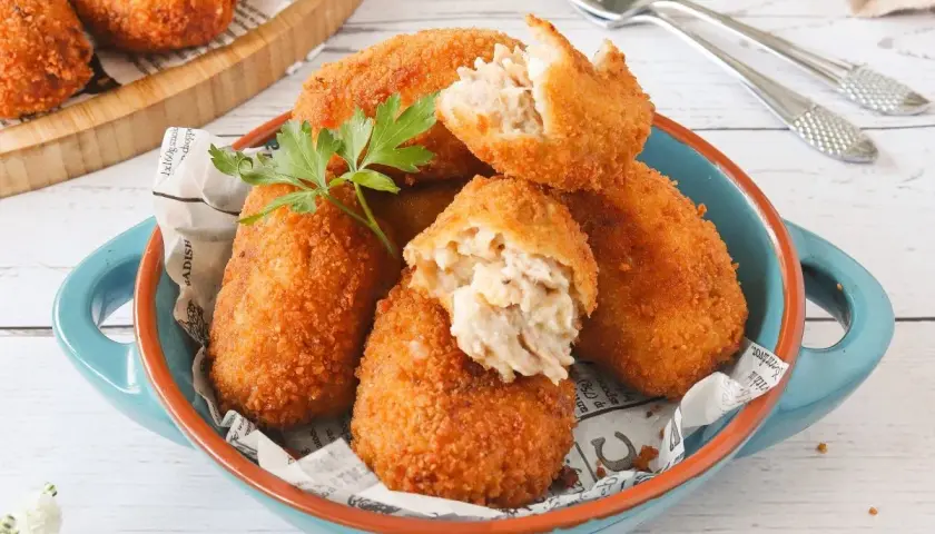 Croquetas de atún doradas y crujientes, servidas en un cuenco azul. Una está partida, mostrando su cremoso interior.