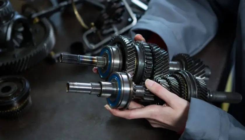 Dłonie trzymające wałek z zębatkami i łożyskami, na tle innych części mechanicznych.