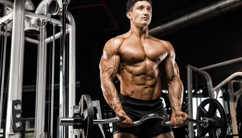 Mężczyzna z wyrzeźbionym ciałem wykonuje ćwiczenia na biceps z łamaną sztangą w siłowni.