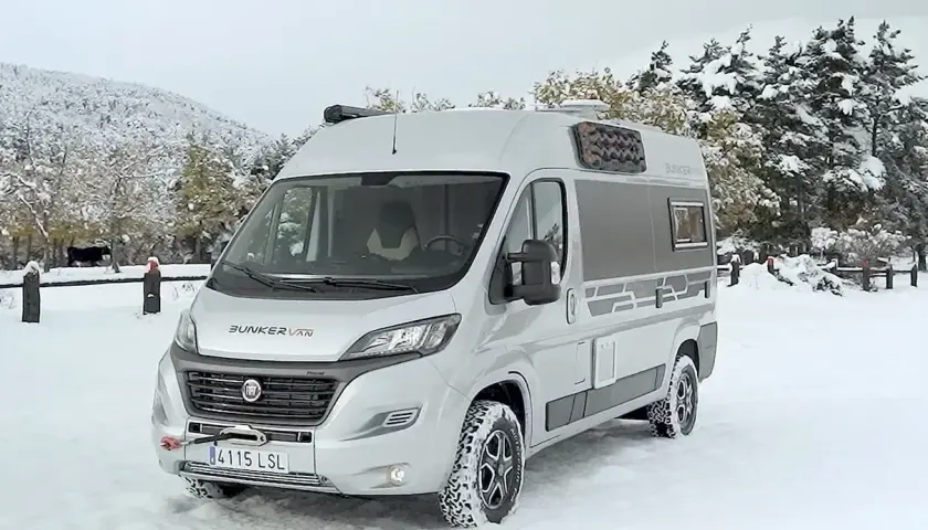 Camping-car 4x4 argenté garé dans la neige, avec des arbres enneigés en arrière-plan.