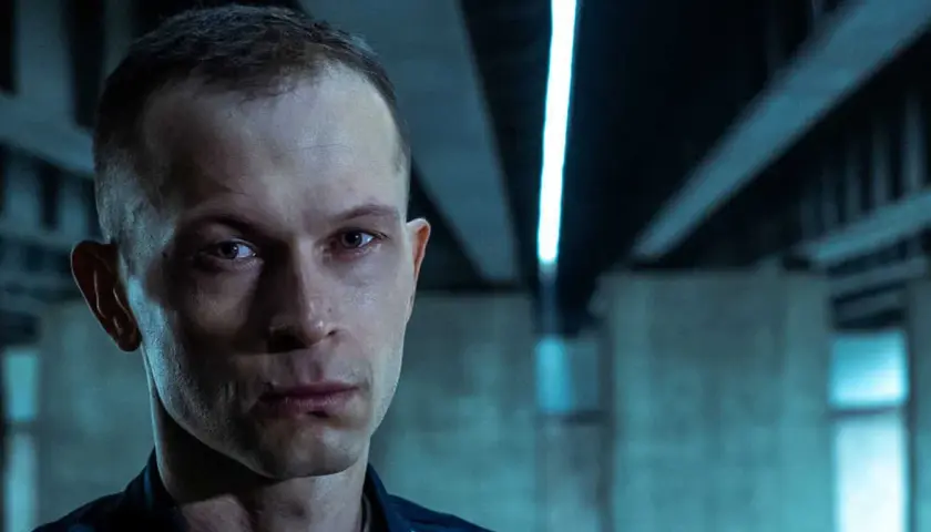Zbliżenie na twarz mężczyzny z filmu "Tomasz Komenda". Jego spojrzenie jest intensywne, a tło sugeruje mroczne otoczenie.