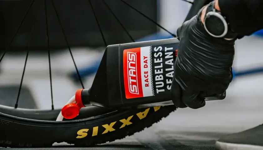 Gloved hands pour Stans Race Day tubeless sealant into a Maxxis tire, demonstrating how to add sealant to tubeless tires for a puncture-free ride.
