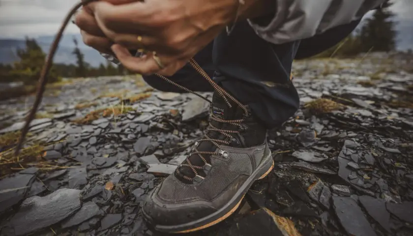 Ręce zawiązują sznurówki wysokich butów trekkingowych na kamienistym, wilgotnym podłożu.