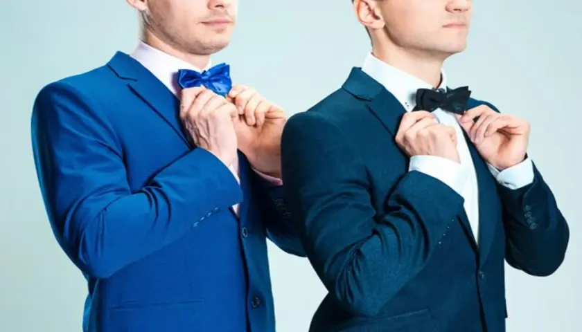 Dwóch mężczyzn w eleganckich garniturach, jeden w niebieskim, drugi w granatowym, poprawia muszki. Jaka mucha do granatowego garnituru?