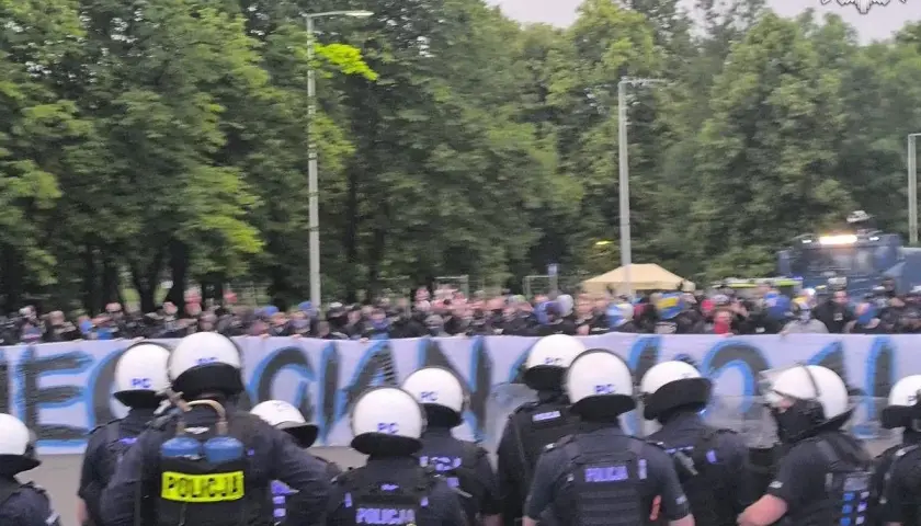 Policja w pełnym rynsztunku stoi przed grupą pseudokibiców, którzy trzymają transparent.
