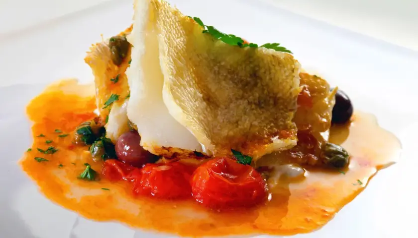Delicioso bacalao encebollado con tomates, aceitunas y alcaparras en una salsa naranja vibrante.