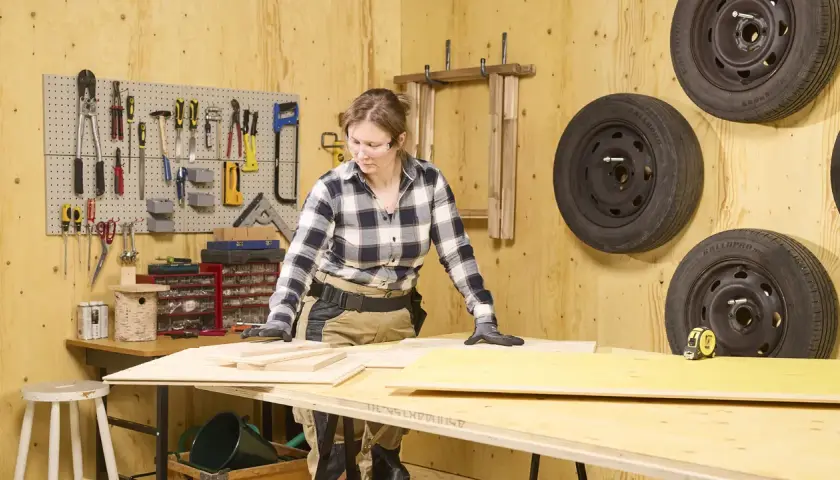 En kvinna arbetar med plywood utomhus i en verkstad. Verktyg hänger på väggen, och däck är upphängda.