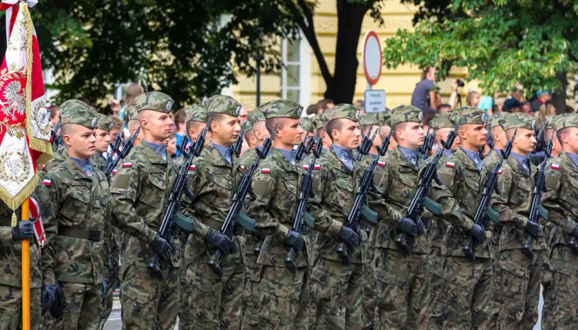 Żołnierze w mundurach i beretach stoją w zwartym szyku, trzymając broń. W tle widać drzewa i budynki, a także flagę. To widok stanowisk wojskowych podczas parady.
