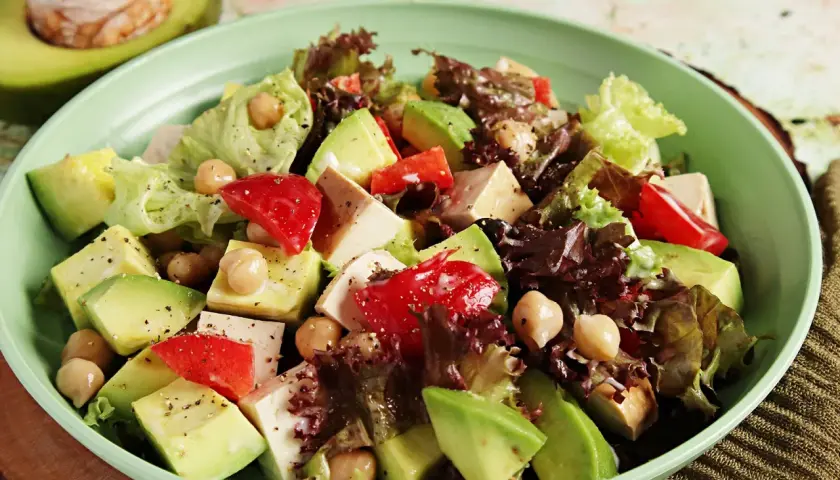 Frischer veganer Salat mit Avocado, Kichererbsen, Tofu und roten Paprikastücken in einer hellgrünen Schüssel.