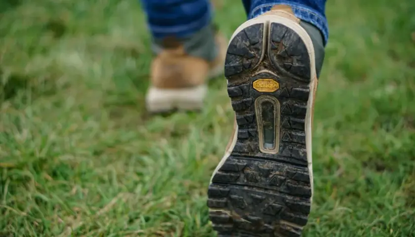 Osoba w dżinsach i butach trekkingowych stawia kroki na trawie. Czy buty trekkingowe powinny być większe? Zobacz podeszwę Vibram.