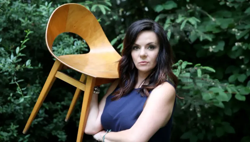 Krystyna Łuczak-Surówka, w wieku, z dumą prezentuje designerskie krzesło na tle zieleni.