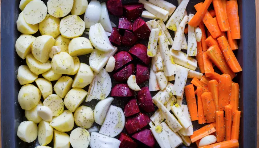 En ugnsplåt med förberedda rotfrukter i ugn: potatis, lök, rödbetor, palsternacka och morötter, kryddade och oljade.