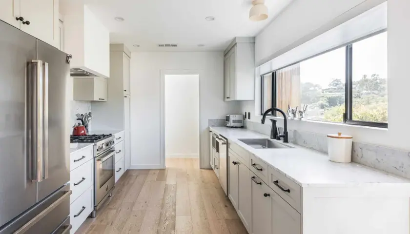 A bright, modern galley kitchen with white cabinets, stainless steel appliances, and a large window overlooking greenery.