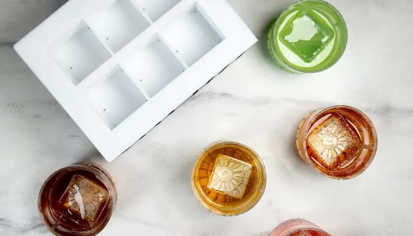 En vit form för klar is bredvid glas med drinkar. Varje drink har en stor, klar isbit med ett mönster.