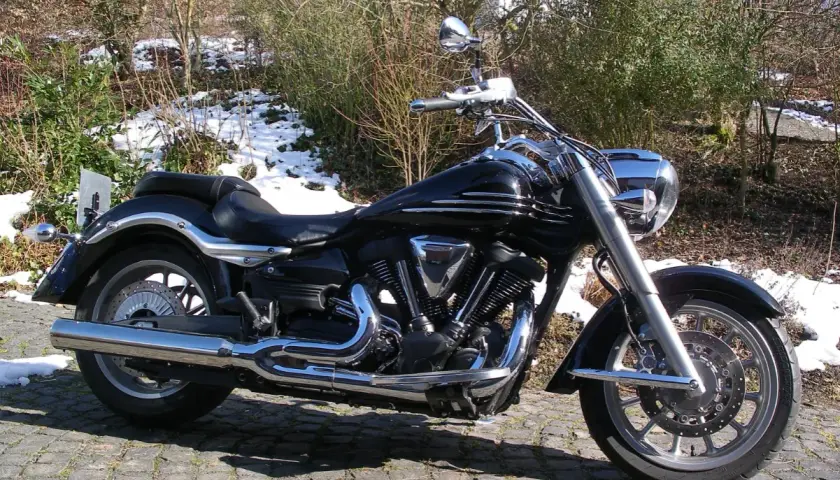 Błyszczący czarny motocykl Yamaha Raider 1900, z chromowanymi detalami, zaparkowany na bruku. W tle zima i krzewy.