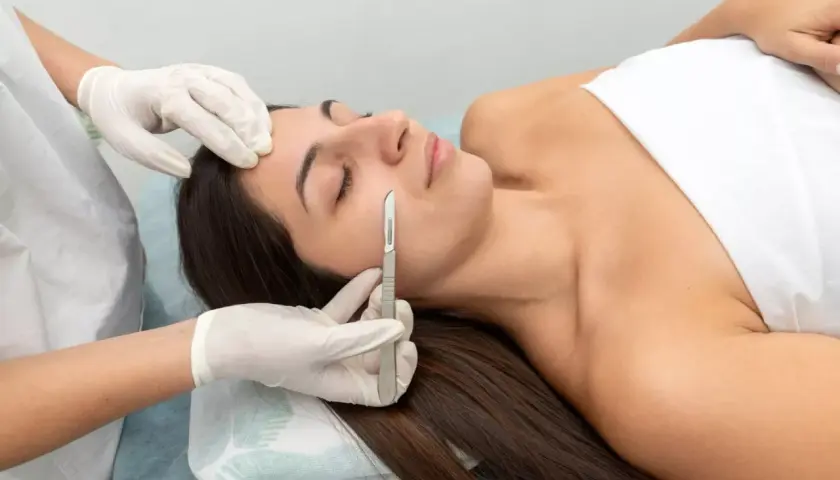 Una esteticista realiza un dermaplaning facial a una mujer, eliminando vello fino y células muertas con un bisturí.