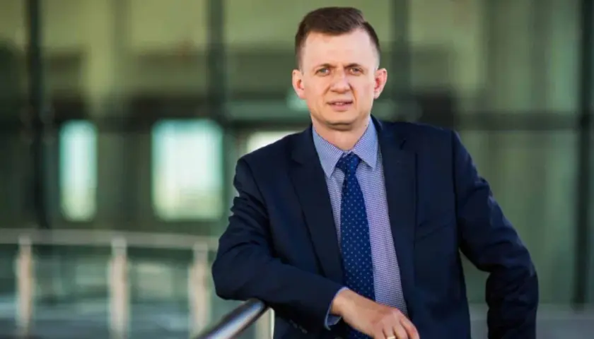 Mężczyzna w garniturze i krawacie opiera się o barierkę na tle nowoczesnego budynku.