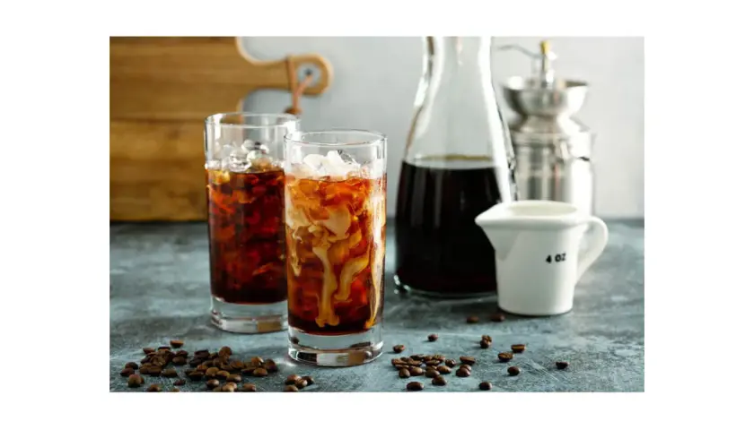 Dwa szklanki orzeźwiającego cold brew z lodem i mlekiem, obok dzbanek z kawą i młynek. Idealne na gorące dni, przygotowane z myślą o miłośnikach kawy.