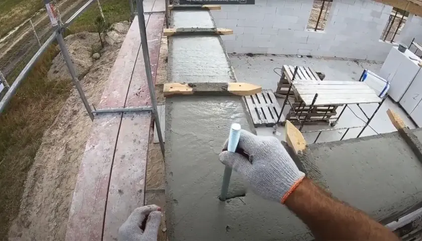 Ręka w rękawicy wkręca pręt w świeży beton. To mocowanie murłaty bez wieńca, kluczowe dla stabilności konstrukcji.