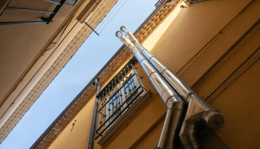 Dwa błyszczące, metalowe rury tworzą komin systemowy z wentylacją, biegnący wzdłuż żółtej ściany budynku.