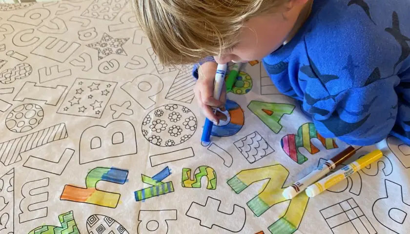 Niño rubio colorea un mantel con letras y dibujos. El **cuadrado para colorear** se llena de vida con sus rotuladores.