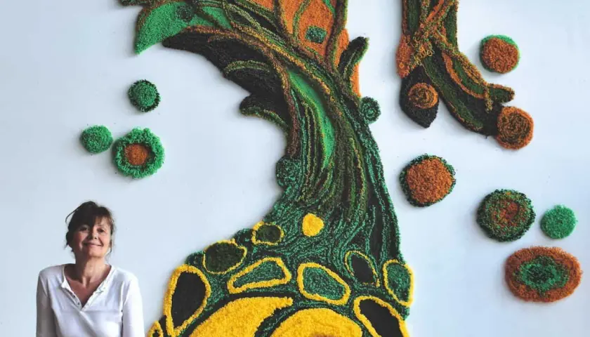 Une designer textile pose devant sa création murale en relief, un arbre aux couleurs vives.