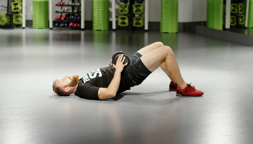 Mężczyzna z brodą wykonuje ćwiczenie glute bridge z obciążeniem na biodrach.