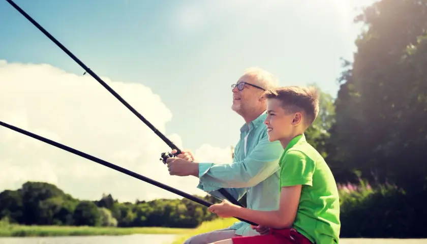 Abuelo y nieto disfrutan de un día de miños pesca, con cañas en mano y sonrisas en el rostro, bajo un cielo azul y soleado.