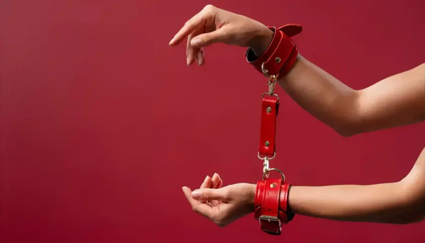 Czerwone kajdanki na nadgarstkach, symbolizujące intymną relację BDSM, na tle bordowej ściany.