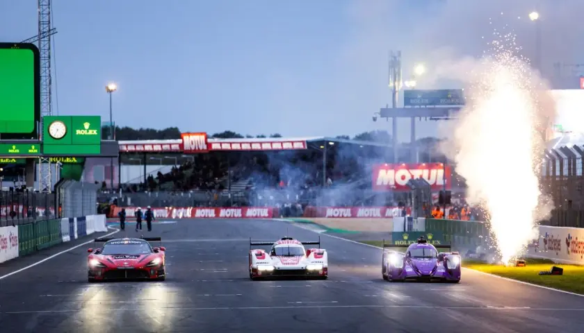Tres coches de carreras compiten en Le Mans, con fuegos artificiales iluminando la pista. El horario de Le Mans se acerca.