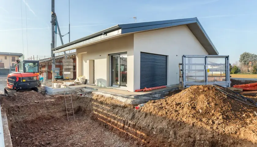Budowa domu: wykop pod fundamenty, koparka i częściowo wykończony budynek.