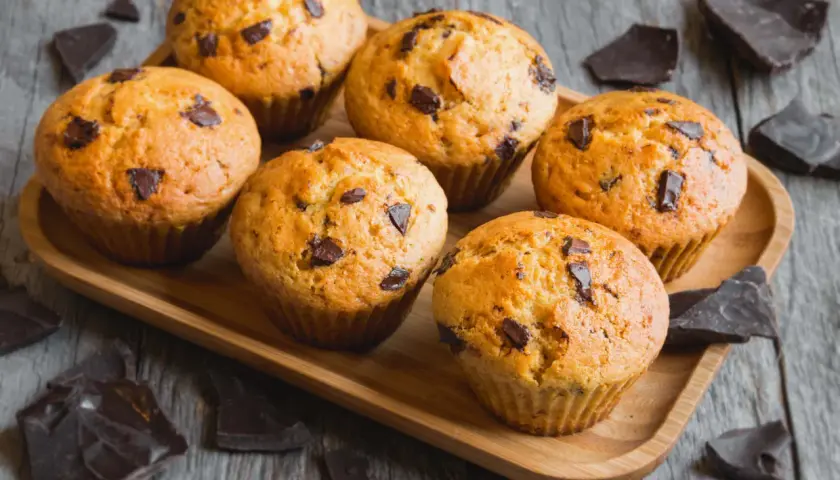 Sei muffin allo yogurt con gocce di cioccolato su un vassoio di legno, circondati da pezzi di cioccolato fondente.