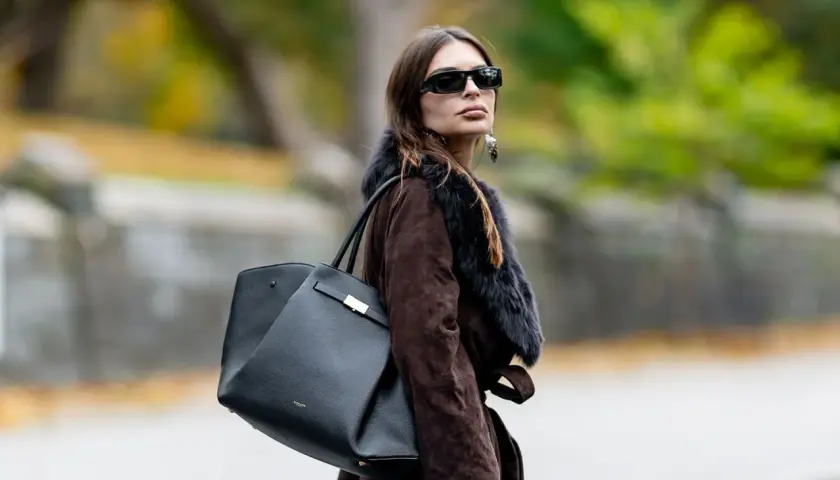Mujer con abrigo marrón y bolso negro de Demellier, gafas de sol y pendientes llamativos.