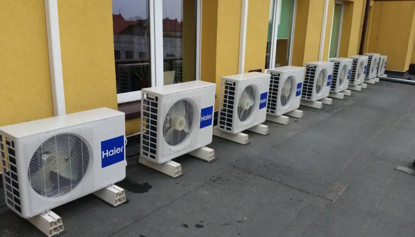 Rząd białych jednostek klimatyzacyjnych Haier na dachu, obok żółtej ściany budynku.