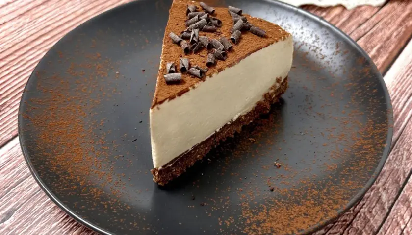 Una fetta di cheesecake al caffè, cosparsa di cacao e riccioli di cioccolato, su un piatto nero.