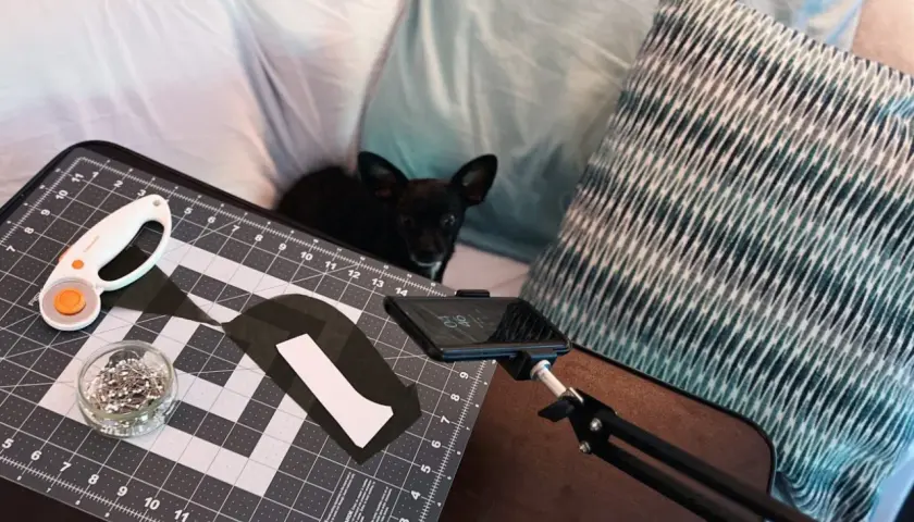 A small black dog watches as someone uses adaptive sewing tools, including a rotary cutter and pins, on a cutting mat.