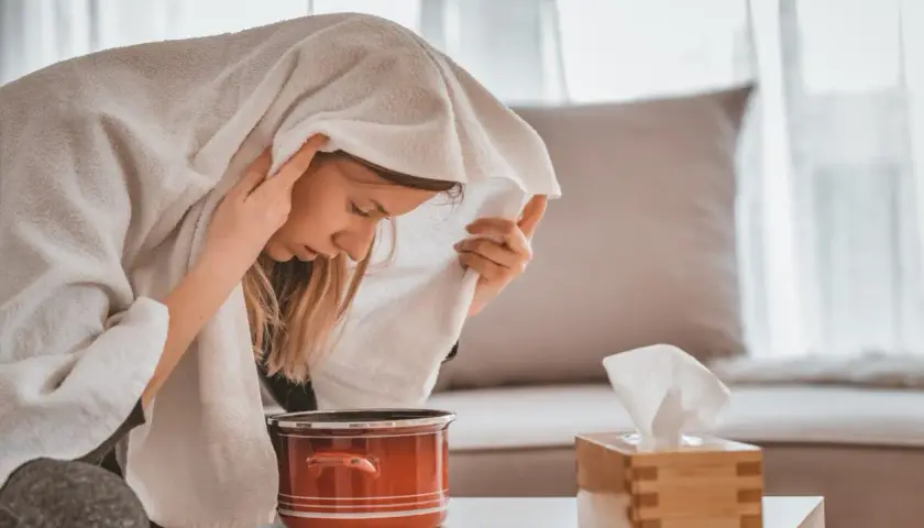 Kobieta wykonująca inhalację parową nad garnkiem z wodą, przykryta ręcznikiem.
