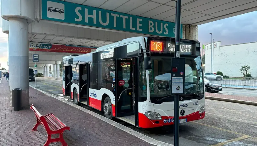 Autobus wahadłowy na lotnisku w Bari, z otwartymi drzwiami, gotowy do odjazdu. Czerwona ławka czeka na pasażerów.