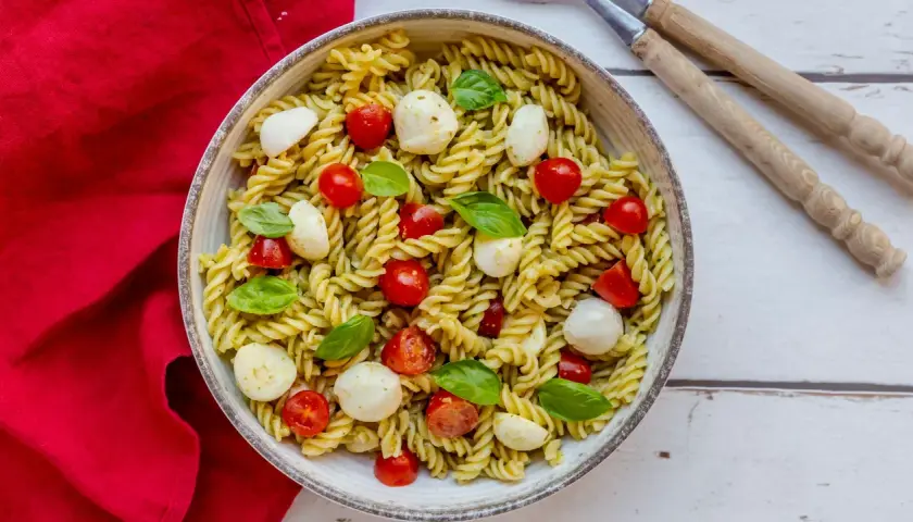 Un piatto invitante di pasta fredda al pesto, arricchito da ciliegini succosi e mozzarelline fresche, guarnito con basilico profumato.