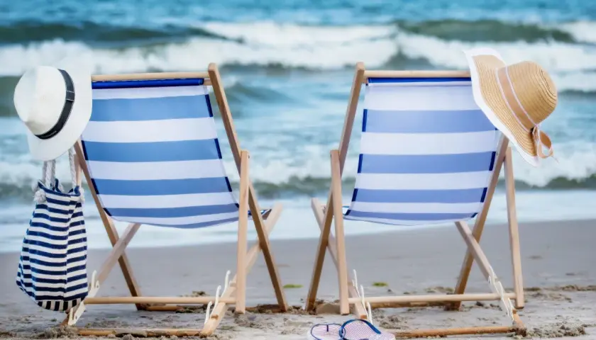Dwa leżaki na plaży, gotowe na idealny weekend nad morzem. W tle fale, a na leżakach kapelusze i torba.