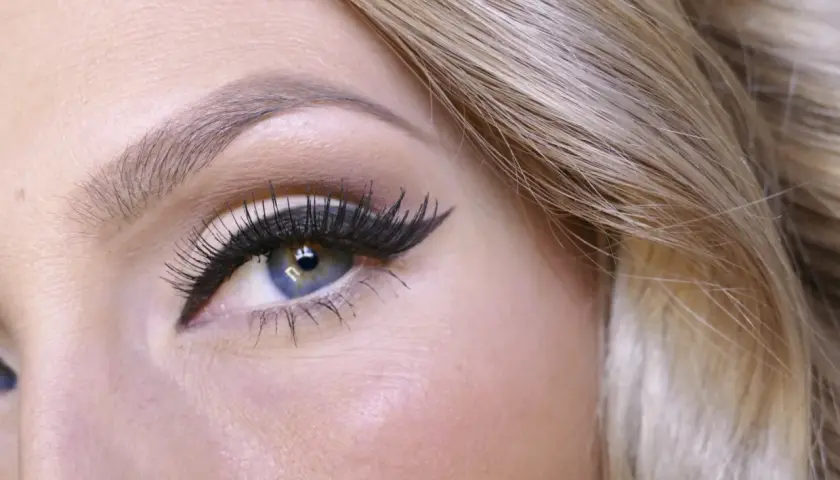 En närbild på ett blått öga med perfekt målad eyeliner och långa fransar. Håret är blont och böljande.