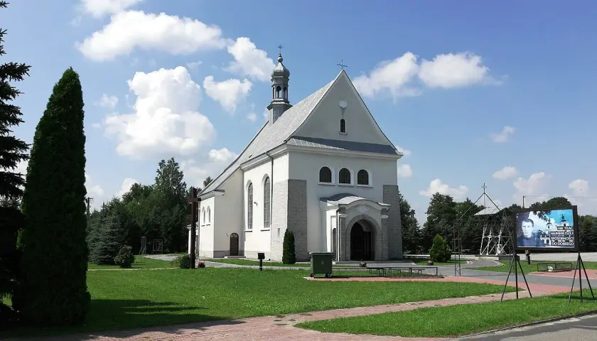 Kościół w Dulczy Wielkiej, z wieżą i krzyżem, na tle błękitnego nieba z chmurami. Obok tablica z wizerunkiem młodego mężczyzny.