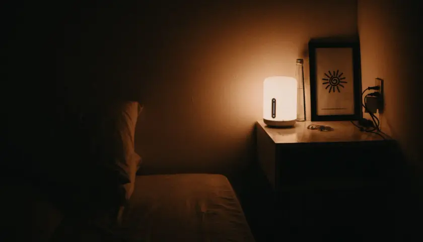 Łóżko z poduszką w półmroku. Lampka nocna rozprasza mrok, łagodząc objawy nyktofobii. Na stoliku butelka i obrazek.