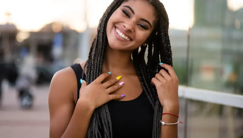 Joven sonriente con trenzas africanas, uñas de colores y un brazalete rojo.