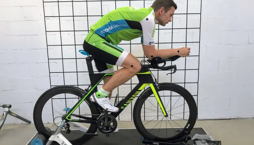 Mężczyzna w stroju kolarskim na rowerze stacjonarnym podczas sesji bike fitting. Punkty pomiarowe na ciele i rowerze.